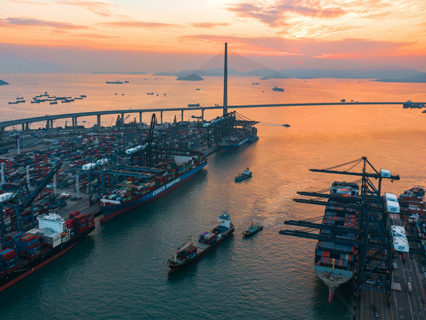Container port with ships and cranes at sunset near a bridge.